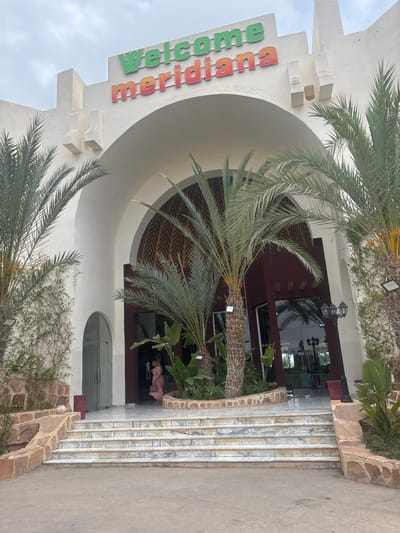 A wide-angle, eye-level shot shows the entrance to the Meridian Beach Resort in Taguermess, Tunisia. The building's facade is white, with a large arched entrance. Above the arch, the words "Welcome Meridiana" are displayed in colorful, raised lettering. Green letters spell out "Welcome" and red and orange letters spell out "Meridiana." Two large palm trees flank the entrance. The steps leading up to the entrance are made of white marble with grey veins. A woman in a pink dress walks up the stairs towards the entrance on the right side of the image. The sky is overcast.