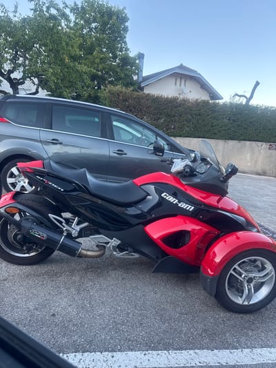 A red and black Can-Am Spyder three-wheeled motorcycle is parked on an asphalt surface. The motorcycle is positioned to the right of a dark grey minivan. In the background, there is a hedge, a house with a peaked roof and a chimney, and trees with green foliage. The sky is clear and blue. The scene appears to be in a residential area, likely in Lavans-lès-Saint-Claude, France, given the presence of a French street sign reading "PLACE" in the background.
