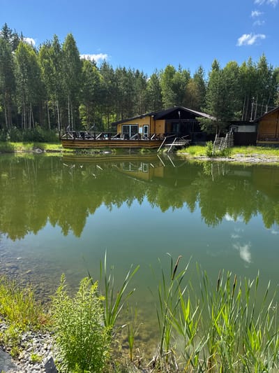 The image depicts a tranquil scene featuring a wooden house with a deck overlooking a calm body of water, likely a lake or pond, situated in the city of Vorsma, Russia. The house, constructed with natural wood, has a modern design with large windows and a dark-pitched roof. A wooden deck extends from the house, furnished with dark chairs and tables, suggesting an outdoor seating area. The deck is bordered by a wooden railing. Surrounding the house is a dense forest of tall, slender trees, predominantly coniferous with some deciduous varieties visible. The water in the foreground reflects the sky, trees, and the house, creating a mirrored image of the landscape. Reeds and other aquatic plants grow along the edge of the water, with some also present in the shallow, rocky area visible on the left. The sky is a clear blue with a few scattered white clouds. The overall impression is one of a peaceful, natural setting, possibly a resort or private residence, designed to harmonize with its environment.