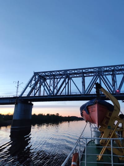 The image shows a view from a boat on a river, with a large steel truss bridge spanning the waterway. The bridge has a concrete pier on the left side of the frame and an electrical pole on the deck, and appears to be for rail use. An orange lifeboat, partly covered with a blue tarp, hangs from a davit on the deck of the boat. The sky is clear with a gradient from light blue near the top of the frame to orange near the horizon. Trees line the shore in the background. The location is likely Нифантово, Russia, considering the style of the bridge and the presence of Russian text on the lifeboat.