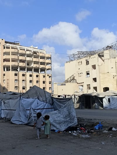 Two young girls walk hand-in-hand in front of a row of makeshift tents. The girl on the left wears a grey t-shirt and patterned leggings, while the girl on the right wears a light green dress. Behind them, damaged buildings stand against a blue sky with scattered white clouds. The building on the left is a tall, multi-story structure with many empty, open window frames and balconies, appearing to be a hotel as indicated by the vertical "HOTEL" text visible on its side. The building on the right is also damaged, with its roof partially collapsed and exposing structural metalwork. The tents in the foreground are made of grey and blue tarpaulin, suggesting a temporary shelter situation. Scattered debris, including plastic bags and other refuse, litters the ground in front of the tents and between the tents and the damaged buildings. The scene appears to be set during the daytime, with natural light illuminating the area. The overall impression is one of devastation and hardship, likely due to conflict or disaster.