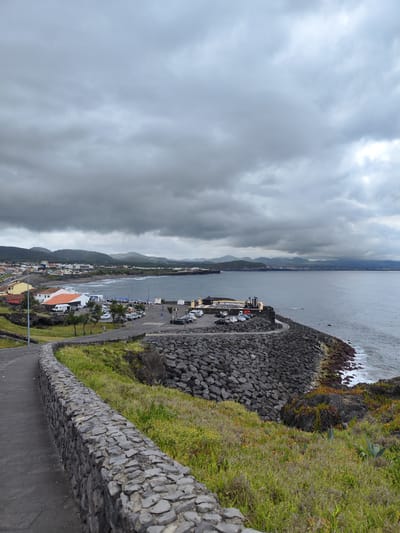 The image shows a coastal view of Ribeira Grande (Matriz), Portugal, under a cloudy sky. In the foreground, a paved pathway curves to the left, bordered by a low, stone wall constructed from rough-hewn rocks. Beyond the wall, grassy slopes rise, interspersed with dark volcanic rocks. To the right, a massive, gray rock breakwater extends into the ocean, with waves crashing against its base and some red and green vegetation growing at its edge.

Further into the scene, a town lines the coast, with buildings of various colors, including white, yellow, and orange roofs. A stretch of beach with gentle waves is visible. Beyond the town, a line of hills forms the backdrop. A parking lot is situated near the coast, with several cars parked. The ocean is a deep blue-gray, reflecting the overcast sky. The overall atmosphere is one of a moderately populated coastal town on a rather gloomy day.