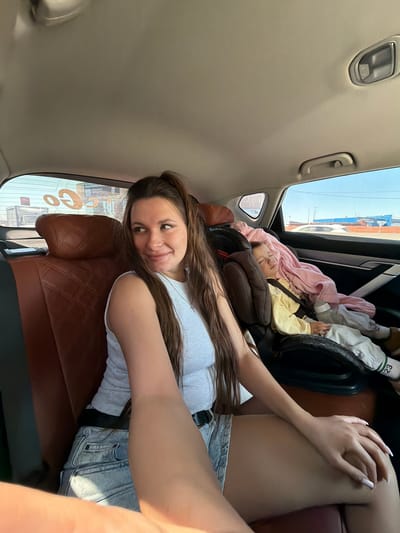 The image is a selfie taken inside the rear cabin of a car. An adult female is seated in the left rear passenger seat, angled slightly towards the camera, with her head turned to look towards her right, away from the camera. She has long, dark brown hair tied in a ponytail, wears a light grey sleeveless ribbed top, light-wash denim shorts with a black belt, and a silver necklace with a cross pendant. Her right arm is extended, likely holding the camera device, and her left hand rests on her left thigh. Her fingernails are long and manicured with light-colored polish.

To her right, in the adjacent right rear passenger seat, a young child (likely a toddler or preschooler) is sleeping in a brown child safety seat, secured by a harness. The child wears a light yellow long-sleeved shirt, light-colored pants with text including "123" on the left leg, and brown shoes. A pink blanket is draped over the child's lap and torso.

The car's interior features brown leather seats with a diamond-quilted pattern. The ceiling is a light beige or grey fabric, showing grab handles and an overhead light fixture.

Through the car windows, an exterior environment is visible. The rear window reflects an orange "GO" graphic (reversed) and displays faint, reversed Cyrillic text "СКИДКИ ПО" (Skidki Po - "Discounts on"), indicative of a sign in a Russian-speaking region. The side windows reveal a bright blue sky, power lines, and a road with other vehicles. A white car and a large, blue, industrial-style building are visible in the distance.

The scene depicts individuals in transit within a vehicle in an urban or suburban area of Trekhgorka, Russia, as evidenced by the Cyrillic script in the background.