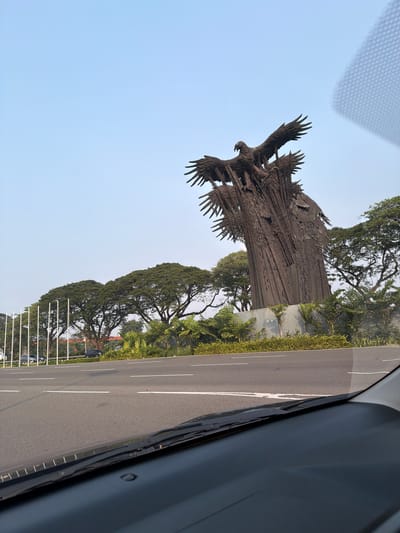 This image, taken from inside a vehicle, shows a large, abstract sculpture of eagles with outstretched wings. The sculpture is made of dark, textured metal and appears to be mounted on a concrete base. It is situated outdoors, next to a road with a grassy median. Tall trees with dense green foliage line the road, creating a lush, natural setting. The sky is clear and light blue, suggesting a bright day. The perspective of the photo, with the road and sculpture appearing on their sides, indicates that the image was likely taken from a car window, possibly during a drive. The angle also suggests the car is moving, as the side mirror is visible at the edge of the frame. The scene appears to be a public space, possibly a roadside attraction or memorial, given the prominent sculpture and well-maintained road. There are no people visible in the frame. The overall mood is one of grandeur and nature, with the imposing sculpture set against the backdrop of mature trees and a clear sky.