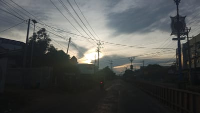 The image depicts a street scene in Geulanggang Teungoh, Indonesia, during what appears to be twilight or early morning. Overhead power lines crisscross the sky, supported by poles. The sun is partially visible behind clouds, casting a glow and shadows across the scene. On the left side of the road, there are buildings and dense foliage. A road stretches into the distance, with a railing visible on the right side. A lone motorcycle is in motion on the road. Streetlights, some with ornate decorative elements, are present along the street. The overall atmosphere is somewhat dim, with the sky dominated by cloudy textures.