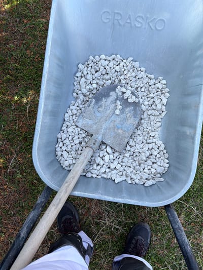 An overhead view captures a galvanized metal wheelbarrow, embossed with the name "GRASKO," positioned outdoors on a patch of sparse grass in Inovo, Bulgaria. The wheelbarrow's pan contains an accumulation of light grey, angular gravel, over which a well-worn metal shovel with a wooden handle rests blade-down. The shovel blade exhibits traces of blue-grey residue. At the bottom of the frame, the lower legs and feet of an individual are visible, wearing black work boots with red laces and white trousers featuring dark-colored accents on the shins.