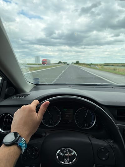 The image is a first-person perspective shot from inside a car, as indicated by the steering wheel, dashboard with a speedometer and other indicators, and the driver's hand on the wheel. The car is on a highway, with other vehicles visible in the distance, including a red truck. The road appears to be a multi-lane highway with grassy shoulders and a cloudy sky above. The dashboard displays a speed of approximately 130 km/h. The driver is wearing a smartwatch. The scene appears to be on a highway in or near Zavoy, Bulgaria, given the context. The environment is that of a typical highway drive.
