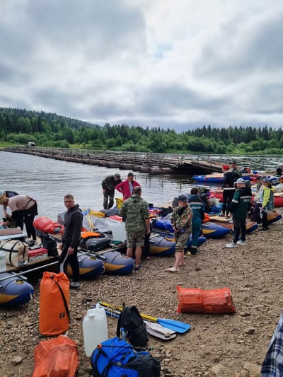 The image depicts a group of people gathered on a rocky shore next to a river, with a makeshift pontoon bridge extending into the water in the background. The scene appears to be a staging area for an outdoor activity, likely involving rafts or kayaks, as several inflatable boats are visible both on the shore and in the water. People are engaged in various activities, including loading and organizing gear, conversing, and standing by.
The environment consists of a wide river with moderately flowing water, bordered by a dense forest of coniferous and deciduous trees stretching up a hillside. The sky is overcast with clouds. The shore is covered in gravel and small stones, with scattered pieces of debris.
Essential details include:
* **People:** Approximately 15-20 individuals are visible, dressed in casual outdoor attire, some in what appear to be specialized gear or uniforms (one person wears a jacket with "ЭКОС" written on the back). They are of varying ages and genders.
* **Objects:** Numerous inflatable rafts and kayaks are present, in various colors including blue, yellow, and red. There are also bags, duffel bags, dry bags (some in bright orange), a white water container, a blue and gray paddle, and possibly fishing equipment or poles. In the background, the pontoon bridge is constructed from logs and appears to be a temporary structure.
* **Actions:** People are observed standing, bending over to pack or unpack items, and looking in different directions. Some are standing in small groups, suggesting conversation or coordination.
* **Environment:** The location is a natural setting with a river, rocky shore, and forested hills. The weather is cloudy, indicating a cool or damp day.
While the image doesn't explicitly provide information to pinpoint the exact location within Вишерогорск, Russia, the natural landscape (river, forest, rocky shore) is consistent with a recreational or expedition launch point in such an area. The pontoon bridge suggests a crossing point that might be used for access to remote areas or for specific events.