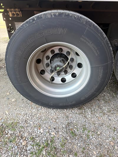 This is a close-up, eye-level, exterior, medium shot of a large truck tire mounted on a steel rim. The tire is black and shows prominent lettering which reads "DOUBLE COIN ALL STEEL RADIAL" on the left side and "295/75R22.5 144/141L 14PR" on the right side. The rim is silver and has multiple lug nuts. A white wire is attached to a green valve stem on the wheel. The background consists of gravel and some patches of green grass. The truck's undercarriage is visible in the upper portion of the image. There are no people or animals present. The location is not specified.