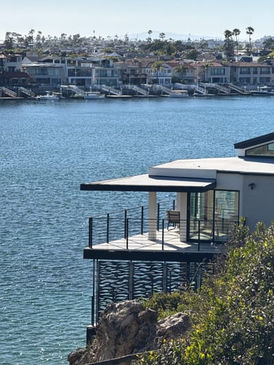 The image depicts a waterfront scene in Newport Beach, California, United States. The foreground features a modern house perched on a rocky outcrop. This house has a deck with dark railings and a geometric patterned screen below. A chair is visible on the deck. The house appears to be built into the cliff, surrounded by greenery. In the background, across the water, is a row of houses along the waterfront, each with docks. Beyond these houses, the skyline is punctuated by palm trees. The water in the foreground is calm and reflects the sky. The day appears bright and sunny.