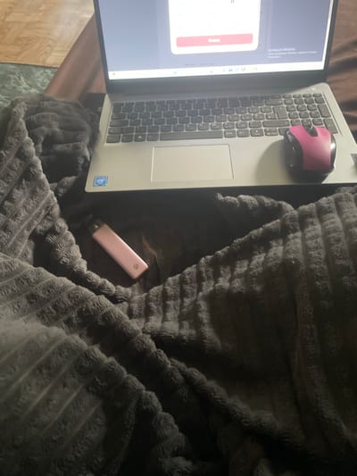 The image shows a pink vape device resting on a dark gray, plush blanket. A laptop computer is partially visible in the background, with a pink computer mouse positioned on its keyboard. The laptop screen displays text and a red button. The scene appears to be indoors, possibly in a living space. There is no discernible connection to the city of Линёвка, Russia, as no geographical markers or specific environmental details indicate this location.