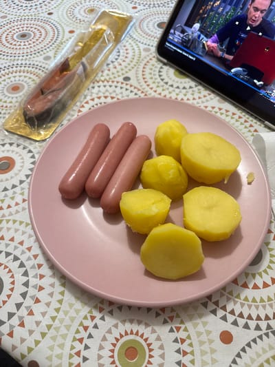 The image shows a plate of food on a patterned tablecloth. The plate contains several boiled potato halves and three uncooked sausages. To the left of the plate is a foil-wrapped packet containing what appears to be fish. In the background, a tablet screen displays a video of a man speaking. The overall setting suggests a casual meal, possibly at home. The lighting appears to be indoor. There is no visible text in the image that provides further context.
