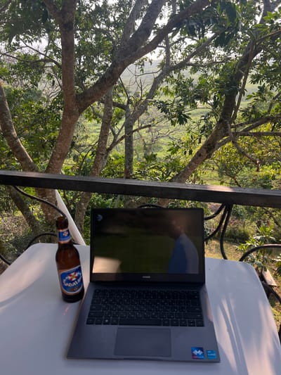 The image depicts a laptop and an open bottle of Salvavida beer on a white table against a backdrop of dense green trees in what appears to be a natural setting. The setting suggests a location in Santa Cruz de Yojoa, Honduras, a region known for its natural beauty. The scene is oriented vertically with a part of railing that separates the view of the surrounding and the table.