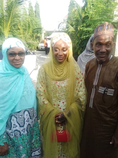 This image depicts an outdoor gathering in Unguwa Uku Kauyen Alu, Nigeria, under bright daylight. Three individuals are prominently featured, posing for the camera.
On the left, an older woman with dark-rimmed glasses wears a light blue hijab and a patterned dress with a green beaded hem, presenting a slight smile. The central figure is a younger woman adorned in an elaborately embroidered light green long dress and a matching layered veil. Her hands display intricate henna designs and are decorated with gold rings. She holds a small, structured red purse with a gold clasp and offers a gentle smile with eyes downcast. To the right, an older man wears a brown traditional garment with white and black embroidery and a multi-colored patterned cap, smiling broadly at the viewer.
A partially visible figure in a grey head covering stands behind the central woman. The background features lush green foliage, including palm trees, a partially visible white car, and distant indistinct figures, some in orange shirts, positioned near a white tent structure on a paved surface.