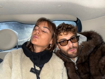 A woman and a man are seated in the back of a car. The woman on the left is wearing a tan fleece jacket with a black hooded scarf, eyes closed, and pursed lips. The man on the right has curly hair, a beard, and is wearing dark glasses. He's sporting a brown fur coat over a gray hooded sweatshirt. The back window behind them appears wet or dirty. The interior of the car is light-colored.