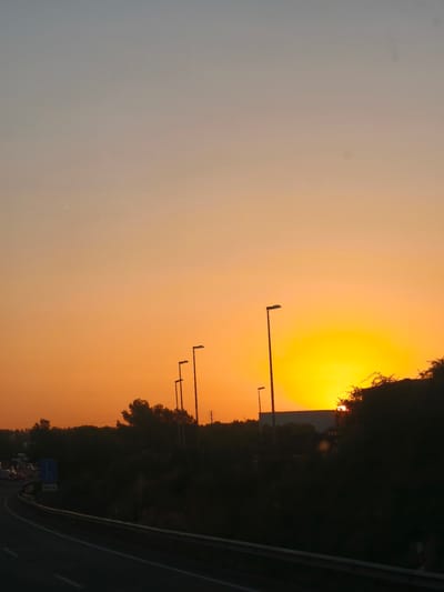 The image captures a sunset scene on a highway in Paterna, Spain. The sky is ablaze with vibrant orange and yellow hues, with the sun partially visible on the horizon. Silhouetted against the sunset are several streetlights, trees, and a highway barrier. In the distance, a traffic jam is evident, with vehicles moving slowly. A blue sign indicating "300m" is also visible on the side of the road. The overall atmosphere is one of a typical evening commute ending with a beautiful natural spectacle.