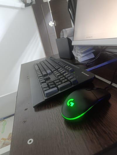 The image shows a close-up, eye-level view of a desk with a computer keyboard and mouse. The keyboard is black, with visible keys and the "Lenovo" logo. The mouse is also black, with a prominent green "G" logo and a green illuminated border. The desk surface is a dark, wood-grain material. In the background, partially visible, is a computer monitor displaying a white screen with a small, faint text that reads "© My Company 2026". To the left of the monitor is a black speaker. To the left of the desk, a white wall is visible, along with some electrical wiring. The image appears to be taken indoors, likely in an office or home setting. There are no people visible in the image. The image does not contain any visual information that would indicate its location in Tursunzoda, Tajikistan.
