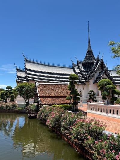 The image depicts a building with a tiered, ornamental roof structure in Bang Pu Subdistrict Municipality, Thailand. The building is adjacent to a body of water. The architectural style is characterized by intricate details and multiple roof levels. The lower part of the building is white with a red brick border. Mature trees and flowering plants border the body of water. The sky is clear blue.