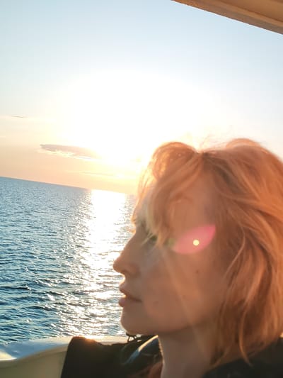 In this close-up, side-profile image, a young person with short, tousled, auburn hair is looking out over a body of water at sunset. The sun is low on the horizon, casting a bright, hazy glow across the sky and reflecting brilliantly on the rippling water. A faint lens flare, appearing as a pinkish circle with a bright dot in the center, is visible on the person's cheek. The person is wearing dark clothing. The water appears calm with gentle waves. The environment suggests an outdoor setting, possibly on a boat or a pier, given the glimpse of a white railing in the foreground. The sky transitions from a pale blue near the top to warm oranges and yellows closer to the horizon. The image is taken from a low angle, with the sky occupying the majority of the frame. The location of Kakkarovo, Russia, is not discernible from the visual information in the image.