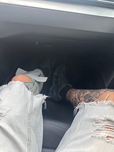 The image depicts a person sitting in what appears to be the interior of a vehicle. The person is wearing ripped light-wash denim shorts and two different pairs of athletic shoes, a dark pair and a light-colored pair, with one shoe having a white paper-like object tucked into it. The individual has a visible tattoo on their right leg. The interior of the vehicle is primarily dark and gray, with a car door panel visible on the left side of the frame. There is no information about the city of Sofronievo, Bulgaria.