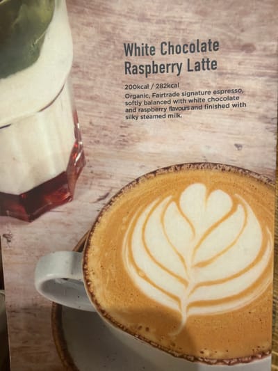 The image shows a promotional shot of a "White Chocolate Raspberry Latte." A frothy, latte art-adorned latte is the main subject of the image, presented in a mug, sitting on a saucer. To the left of the mug is a partially visible glass, likely containing ingredients related to the latte. Text to the left of the drinks advertises the beverage, noting its calorie count, ingredients (including organic, Fairtrade espresso, white chocolate, and raspberry flavors), and texture (silky steamed milk). The scene appears to be set in a cafe or restaurant, potentially in Mole Valley, United Kingdom, given the context.