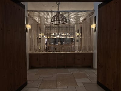 The image shows a dimly lit interior space, possibly a restaurant or bar, viewed through a doorway. The doorway is framed by heavy, dark wooden doors, partially open on either side. Inside, a decorative arrangement of light-colored ropes hangs from the ceiling, partially obscuring the view. A woven, basket-like pendant lamp hangs in the center of the rope arrangement. Below the ropes, a wooden sideboard or counter runs along the back wall. Behind this counter, several people are visible, suggesting a bar or serving area. The floor is tiled with light-colored stone, adding to the warm, muted atmosphere. The location is Ramat Negev Regional Council, Israel.