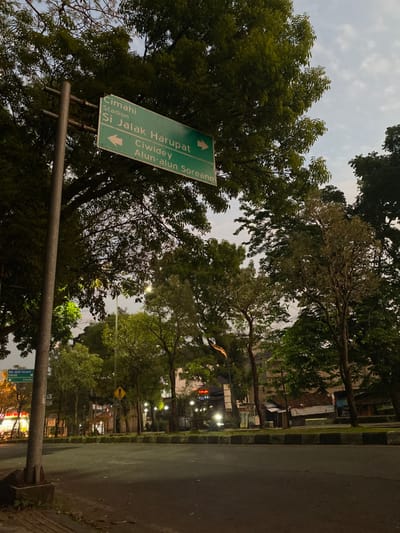 A static, low-light photograph captures an empty urban street scene in Parungserab, Indonesia, likely at dusk or dawn. The foreground features a dark asphalt road, bordered by a black and white checkered concrete curb. To the left, a tall metal pole supports a large, green rectangular road sign. The sign, marked with white text and arrows, indicates "Cimahi" and "Stadion Si Jalak Harupat" with a left-pointing arrow, and "Ciwidey" and "Alun-alun Soreang" with a right-pointing arrow.

Beyond the road, a dense canopy of various trees with green foliage dominates the midground and upper portions of the image, obscuring much of the sky, which appears overcast or twilight-lit. Scattered streetlights are visible among the trees, with some appearing illuminated. In the distance on the right, obscured by foliage, a yellow diamond-shaped sign is partially visible, and structures resembling commercial buildings with illuminated signs, one notably reading "Ayam Goyeng," are discernible. The scene is devoid of people or vehicles.