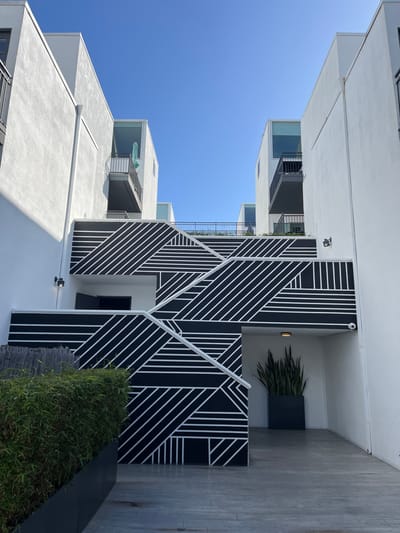 A modern white building reaches into a clear blue sky. A striking black and white geometric pattern adorns the building's facade. Balconies with black railings are visible. A concrete walkway and verdant bushes line the base, with a snake plant in a black planter adding a touch of greenery to the scene. The composition emphasizes the building's angular lines and contemporary design.