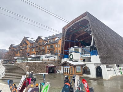 A view of Jay Peak resort. A large A-frame structure covered in wooden shingles towers over a white building with blue accents, marked "Jay Peak." In front, snowboards and skis are stacked. To the left, a multi-story hotel with balconies stands behind a stone wall and a set of stairs. The sky is overcast, and power lines run overhead.