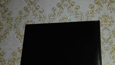 The image shows a close-up of a television screen against a backdrop of ornate wallpaper. The wallpaper features a repeating pattern of swirling floral motifs in gold and cream on a light gray background. The television screen is a solid black rectangle, occupying the majority of the lower portion of the frame. The top edge of the television screen is visible, showing a thin bezel. The lighting appears to be dim, casting a slight shadow on the wallpaper. The location in Занарацкі сельскі Савет, Belarus is not discernible from the image.