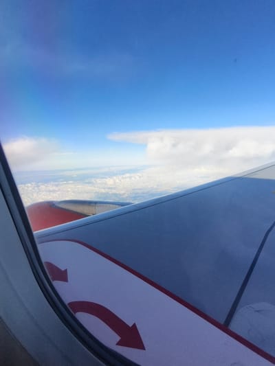 This is a photograph taken from the window of an airplane. The view shows a large airplane wing extending into the frame, with part of an engine visible in red and grey. Below the wing, a vast expanse of white clouds stretches out, with hints of land or a lower layer of clouds visible in the distance. The sky above is a clear, vibrant blue with a few wispy white clouds. On the surface of the wing, there are two red arrows painted on a white background, indicating a direction. The image appears to have been taken during the daytime, possibly from a commercial flight. The specific location of Panschwitz-Kuckau, Germany, cannot be determined from the visual information provided in the image.