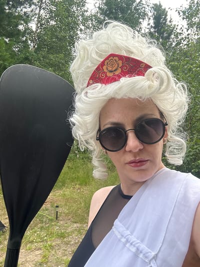 A person, possibly female, stands outdoors in what appears to be a natural setting with trees and grass. They are wearing a white, curly wig with a red and gold floral headband. Round, dark sunglasses are on their face, and their lips are painted with a glossy lipstick. A sheer white garment is draped over their right shoulder. To the left of the person, a portion of a large, black paddle is visible. The background consists of greenery and a cloudy sky. The specific location in Tolbazikha, Russia is not discernible from the image.