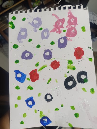 A drawing on a white, perforated notebook page depicts a colorful array of paint splatters. The paint colors include shades of purple, pink, red, blue, black, white and green. The paint is arranged in circular shapes with a white center in several cases, reminiscent of flowers, while other green splatters appear as foliage. The drawing appears to have been executed with a brush and the paint has dripped in some areas. The drawing is held in a person's hand, and in the background, there is a glimpse of a cluttered shelf or storage area. The environment and city of San Juan de Miraflores, Peru, cannot be definitively determined.