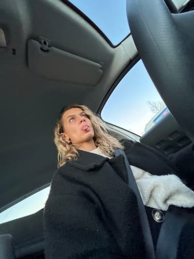 Inside a car, a blonde woman in a black coat is visible. The shot is from a low angle, looking up at the ceiling, the car's sun visor, and through the windshield, which reveals a bright, cloudy sky. The woman's face is tilted upward, with her tongue sticking out. A white, fluffy material is visible inside the coat, possibly a lining or another layer of clothing. A gas station or convenience store is seen through the windshield.