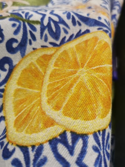 The image is a close-up shot of a piece of fabric with a pattern of orange slices and blue floral designs. The fabric appears to be a tablecloth or similar textile. The orange slices are depicted as round, with a textured interior and a white rind. The blue floral elements are stylized and repetitive, creating a decorative border or background. The lighting is somewhat diffused, and the focus is sharp on the fabric, highlighting the texture of the weave. The background is blurred and dark, suggesting the fabric is draped or positioned against a dark surface. There is no indication of the location being in the city of Pociumbeni, Moldova, or any other specific geographical location. The image solely depicts the fabric itself.