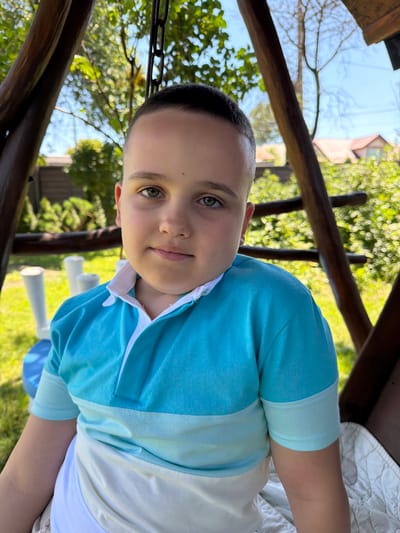 A young boy with short, dark hair and green eyes sits on a swing in an outdoor setting. He is wearing a blue and white horizontally striped polo shirt. The swing appears to be made of wood and has chains. In the background, there is lush green foliage, a wooden fence, and parts of houses with red roofs. The scene is illuminated by natural sunlight, suggesting a daytime setting. The location is identified as Uhornyky, Ukraine.