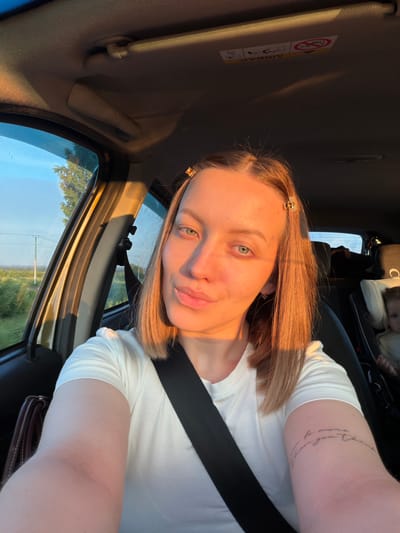 The image shows a woman in the passenger seat of a car, taking a selfie. The sun is shining, casting a warm glow on her face and hair, suggesting it is either sunrise or sunset. She is wearing a white t-shirt and a seatbelt is visible across her chest. A tattoo with script is visible on her upper arm. In the background, a child can be seen in a car seat. Outside the car window, there is a glimpse of green trees and a blue sky, indicating a rural or suburban setting. The car interior includes a sun visor with an airbag warning label. The overall mood is relaxed and casual. The text visible on the sun visor says "AIRBAG" and includes a symbol depicting a person with a deployed airbag.