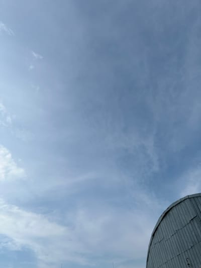 The image captures a portion of a domed structure, likely a roof, against a pale blue sky filled with wispy clouds. The structure appears to be made of metal panels arranged in a grid pattern, with a curved edge visible at the top right of the frame. The sky is overcast with thin, diffused clouds, suggesting a daytime scene with possibly mild weather. There are no people or animals visible in the image. The perspective is from below, looking upwards. The location context is Malyi Oleksyn, Ukraine, though this is not visually evident from the image itself. No text is visible.