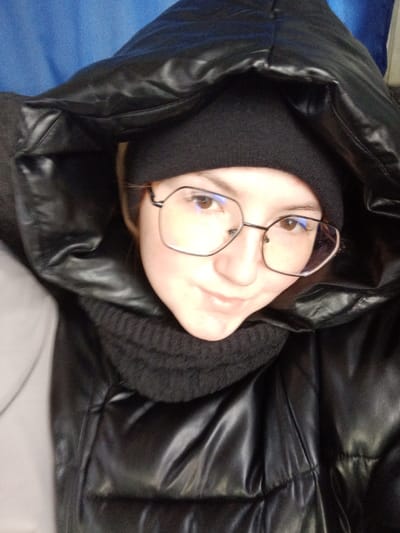 A close-up photograph, captured in the city of Tap'iushka, Russia, features a single female subject positioned centrally and looking directly into the camera. The subject wears a black knit beanie, a black ribbed knit neck warmer, and a black, shiny puffer jacket with an attached hood pulled over her head. She has light-colored skin, brown eyes, and wears hexagonal-shaped glasses with thin, dark frames. Reflections from an unseen light source are visible on her glasses. Her facial expression is neutral to slightly smiling. The background is out of focus, revealing a blurred blue surface in the upper left quadrant and a blurred light gray surface in the lower left, consistent with an indoor or vehicle environment. No other individuals or specific objects are clearly identifiable in the background.