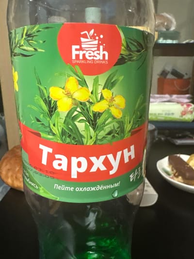 A clear plastic bottle containing a green sparkling beverage is depicted in the city of Cheongju-si, South Korea. The bottle features a green label with a red circular logo at the top, displaying a white drink glass with a straw and leaf, along with the text "Fresh SPARKLING DRINKS." The label illustrates green tarragon leaves and three prominent yellow flowers. A red banner across the lower portion of the label shows the Russian word "Тархун" (Tarkhun) in large white Cyrillic letters, with smaller Korean characters "타르" to its right. Below the banner, the Russian phrase "Пейте охлаждённым!" (Drink chilled!) is visible in green text. Faintly visible under the red banner are the dates "2023.03.10" and "2027.03.09." To the left of the bottle, a light brown, round pastry is partially visible. To the right, on a dark surface, a small white plate with a green rim holds an elongated dessert item partially covered in dark chocolate, alongside a smaller yellow pastry. In the blurry background, a white electronic device, a packet of wipes, and an orange bag are discernible.