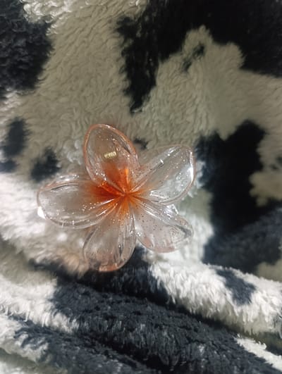 The image shows a plastic, transparent flower-shaped hair clip with orange accents and glitter embedded within the plastic. It rests on a textured, soft fabric patterned with irregular black and white splotches. The background is blurred, focusing attention on the hair clip and its immediate surroundings. There is no indication of the location being San Juan de Miraflores, Peru or any other location. There are no people, actions, or interactions visible.