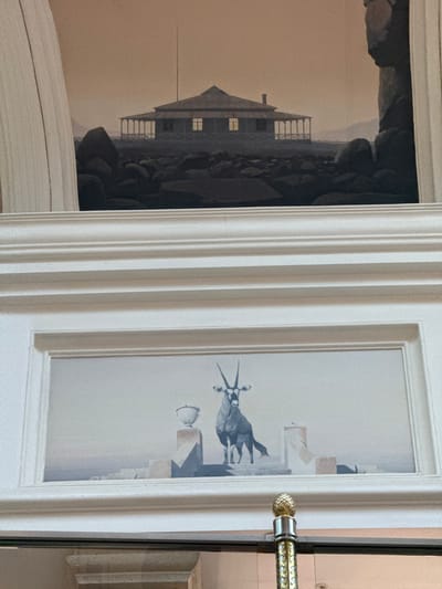 A close-up, vertically-oriented image shows a pair of framed pictures mounted on a wall or possibly a door. The left frame features a dark landscape dominated by a building or structure near a cliff edge, all rendered in shades of grey and brown. The right frame displays a stylized drawing or painting of an oryx or similar horned animal in an arid environment, also in grey tones. The frames are both white with ornate, classical detailing. A door handle, featuring a gold-colored, patterned grip, is affixed to the right side of the frame, suggesting the possibility that the pictures are indeed mounted on a door. The location and city, Milnerton, South Africa are not discernible from the visual elements within the image.