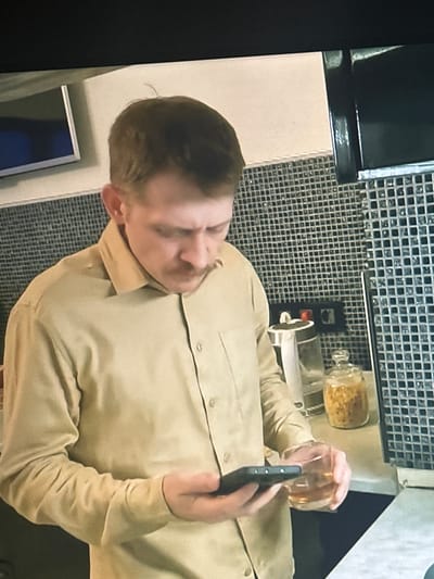 A man with light brown hair and a mustache, wearing a tan button-down shirt, stands in a kitchen. He is holding a smartphone in his left hand and a glass of amber liquid in his right hand. Behind him, a tiled backsplash is visible, along with a glass jar filled with food items. The man is looking down at his phone. The lighting is dim. The location is not specified.