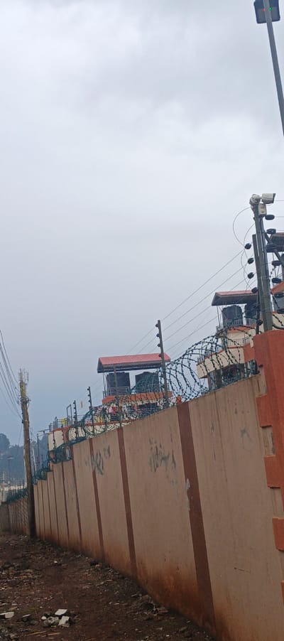 A long beige wall with vertical brown stripes is topped with razor wire and electric fencing under a gray sky. Behind the wall are structures with red roofs and water tanks. A light pole with sensors stands tall on the right. A utility pole with wires runs along the left, and ground cover of dirt and debris is in the foreground. The overall impression is one of security and containment.