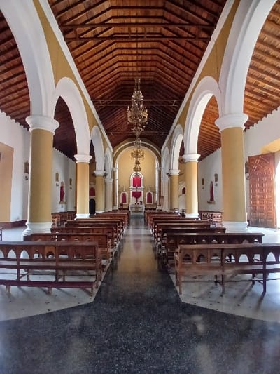 The image displays the interior of a church located in San Joaquín, Venezuela. The perspective is from the rear of the nave, looking forward towards the altar. A central aisle, composed of dark, polished, speckled flooring, extends towards the front. On either side of this aisle, multiple rows of dark wooden pews with carved backrests are arranged, facing the altar.
The architectural elements include tall, ochre-colored cylindrical columns supporting white-faced arches that delineate the nave. The ceiling is a high, pitched wooden structure with visible beams. Two ornate, multi-tiered brass-colored chandeliers hang centrally from the ceiling, decreasing in size towards the front of the church.
At the far end, the altar area is illuminated, featuring a central altar table and prominent red drapery or accents. Along the side walls, between the columns, smaller architectural features or niches with red accents are discernible. On the right side of the image, a large, patterned wooden double door is partially open, allowing natural light to enter the space. No individuals are present in the depicted scene.