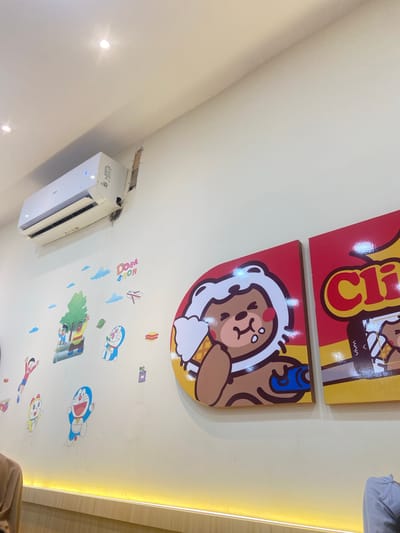 The image shows a wall in what appears to be a cafe or restaurant. The wall is decorated with cartoon characters, including Doraemon and his friends, and two large graphic panels featuring a cartoon bear eating ice cream. An air conditioning unit is mounted on the wall above the decorations. The room is lit by overhead lights and a strip of yellow LED lighting along the bottom of the wall. The general decor suggests a child-friendly or themed establishment, possibly a dessert shop or family restaurant. The specific location within Kembang Harum, Indonesia, is not discernible from the visual information provided in the image.