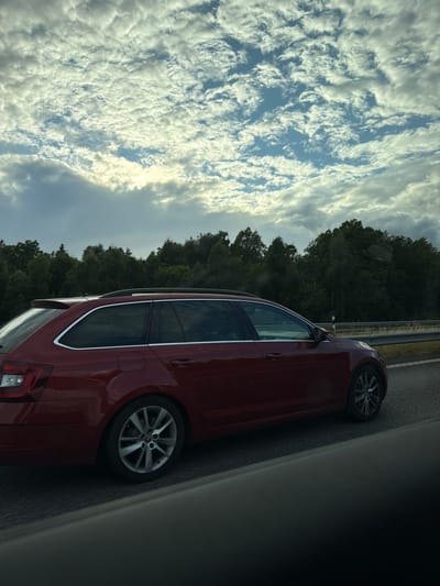 A dark red station wagon is captured in a side profile, traveling on a paved road. The car is a Skoda Octavia, indicated by the logo on the wheel. It has a silver roof rack and silver trim around the windows. The car's rear light cluster and alloy wheels are visible. The background consists of a dense line of dark green trees, suggesting a rural or suburban setting. Above the trees, a sky filled with scattered, puffy white clouds is visible, with patches of blue sky peeking through. The lighting suggests it is daytime, possibly late afternoon, given the soft sunlight filtering through the clouds. The image appears to have been taken from another vehicle, as indicated by the blurred foreground elements. The location is Åskloster, Sweden.