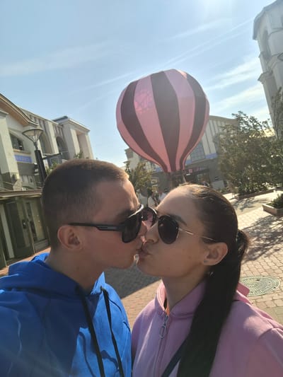 The image presents two individuals, a male and a female, engaged in a kiss in an outdoor pedestrian environment identified as Ingolstadt Village, Ingolstadt, Germany. The male, positioned on the left, wears a blue hooded sweatshirt and black Carrera sunglasses. The female, on the right, wears a pink hooded sweatshirt, gold-framed sunglasses, and has dark hair secured in a low ponytail, displaying a small infinity symbol earring. Their profiles are directed towards each other, with their lips touching. In the background, a large decorative hot air balloon, featuring alternating dark and light pink vertical stripes, is prominently displayed. Surrounding structures include multi-story, light-colored commercial buildings with arched windows; one building clearly shows "BOSS" signage. The ground is paved with light-colored bricks or cobblestones. The scene is illuminated by bright sunlight under a clear blue sky with faint contrails.