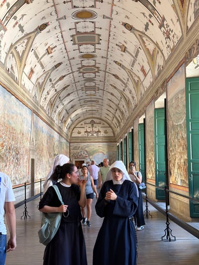 The image depicts an interior walkway within the Royal Site of San Lorenzo de El Escorial in Spain. The walkway is characterized by a vaulted ceiling adorned with intricate frescoes, featuring geometric patterns and various figures. Along the walls, large-scale paintings depict historical scenes, with a prominent mural on the left showing a multitude of people. Green shutters are visible on the right side, set within arched openings.

Several individuals are present in the walkway, appearing to be tourists or visitors. In the foreground, two women are visible. One, on the left, is wearing a black dress and a light blue shoulder bag and is gesturing with her hand. To her right and slightly behind her, a nun in a dark blue habit and white wimple is looking towards the viewer, with her hands clasped. Other people are seen further down the walkway, including a man in a light green shirt and a woman holding a smartphone. The floor is made of wooden planks, and a black metal railing runs along the right side of the walkway, separating it from the wall murals. The lighting suggests it is daytime, with natural light entering from unseen sources.