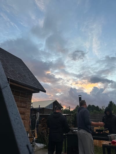 In a rural setting, possibly the Тумілавіцкі сельскі Савет, Belarus, three men are gathered outdoors during twilight. The sky is filled with dramatic clouds, with hints of orange and yellow light breaking through, suggesting either sunset or sunrise. In the foreground to the left, a portion of a dark, angular object, possibly part of a structure or equipment, is visible. Adjacent to it, a wooden building with a dark, shingled roof is prominent.
The three men are positioned around an outdoor cooking area. The man on the far left, wearing a dark hooded jacket and a baseball cap, appears to be tending to a pot or dish. The central figure, a man in a grey hooded sweatshirt and light-colored sweatpants, is turned slightly to his right, gesturing with his hand and looking towards the man on his right. The third man, on the far right, is wearing a dark hooded jacket and is facing the central figure, holding a pan over a heat source. Behind them, a stack of firewood and some construction materials, including blue scaffolding and white string lights, are visible. In the background, the silhouette of trees and another building with a light-colored roof can be seen. The ground appears to be paved or covered in gravel.